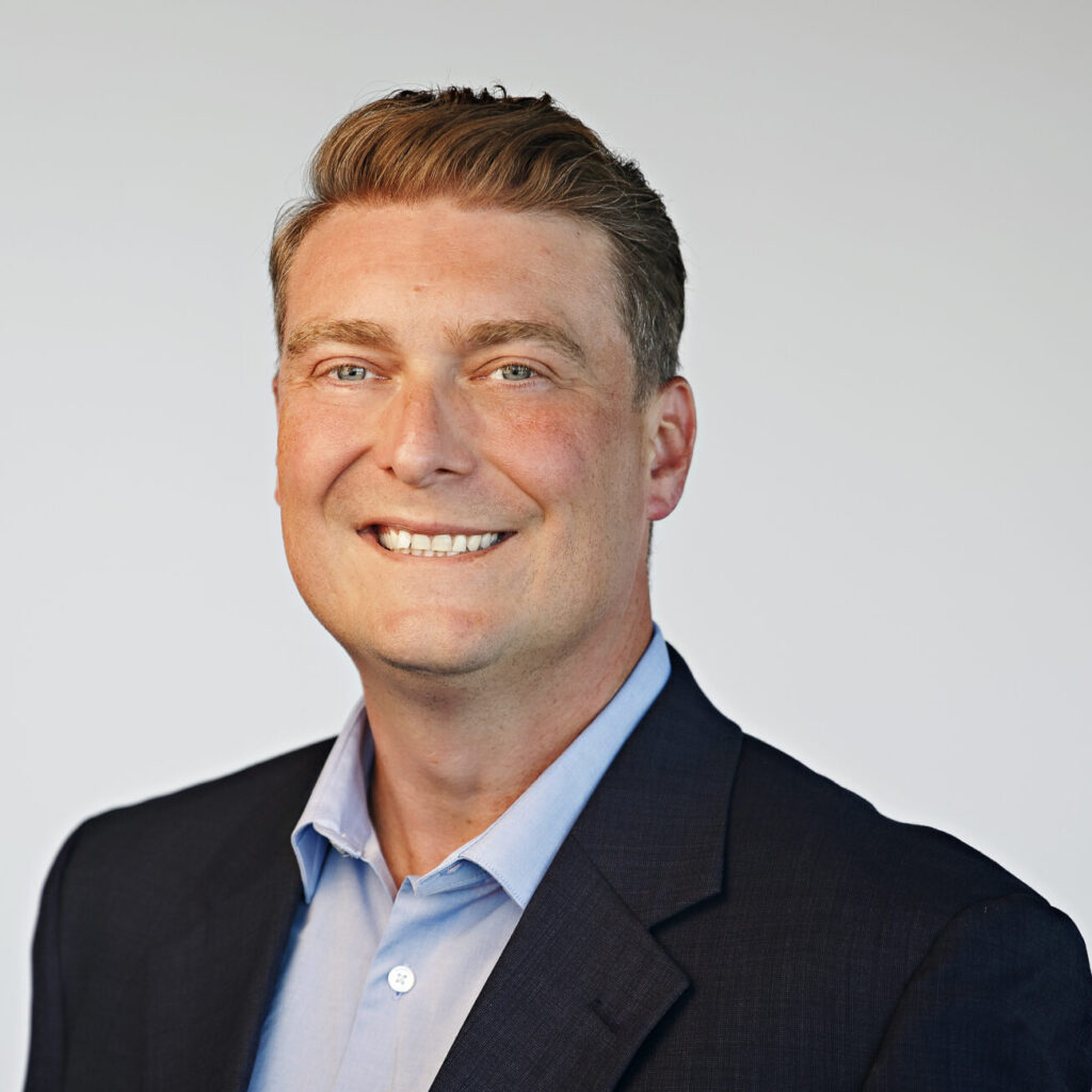 Smiling man in a dark suit jacket and light blue dress shirt, facing forward against a plain light background.