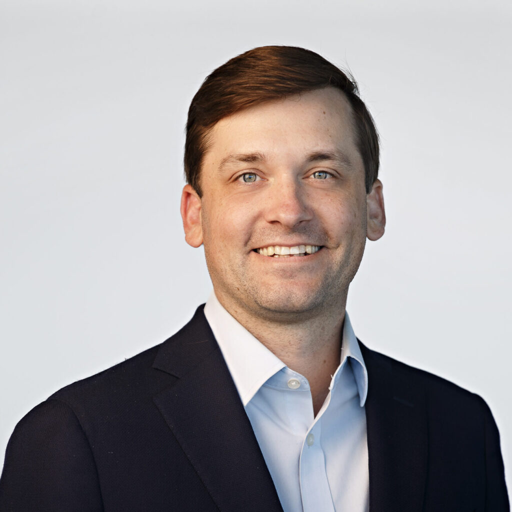 A man in a dark suit jacket and light blue shirt smiles at the camera against a plain light background.