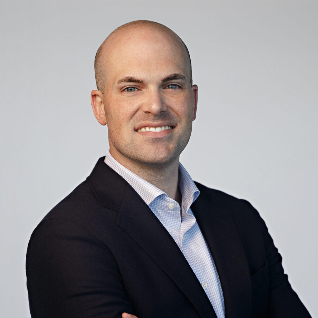A man in a dark suit jacket and light dress shirt stands with arms crossed, smiling at the camera against a plain light background.