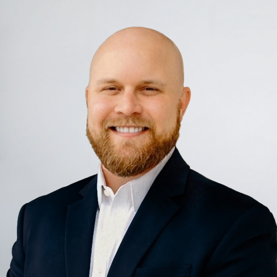 A bald man with a beard, wearing a black suit jacket and white shirt, smiling in front of a plain light background.